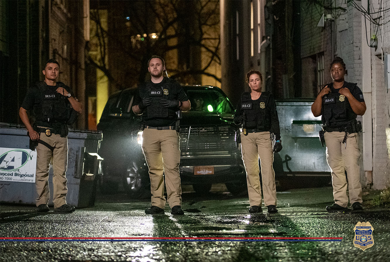 Four IRS-CI officers in tactical gear confidently advancing down a wet alleyway at night, reflecting a sense of readiness and vigilance