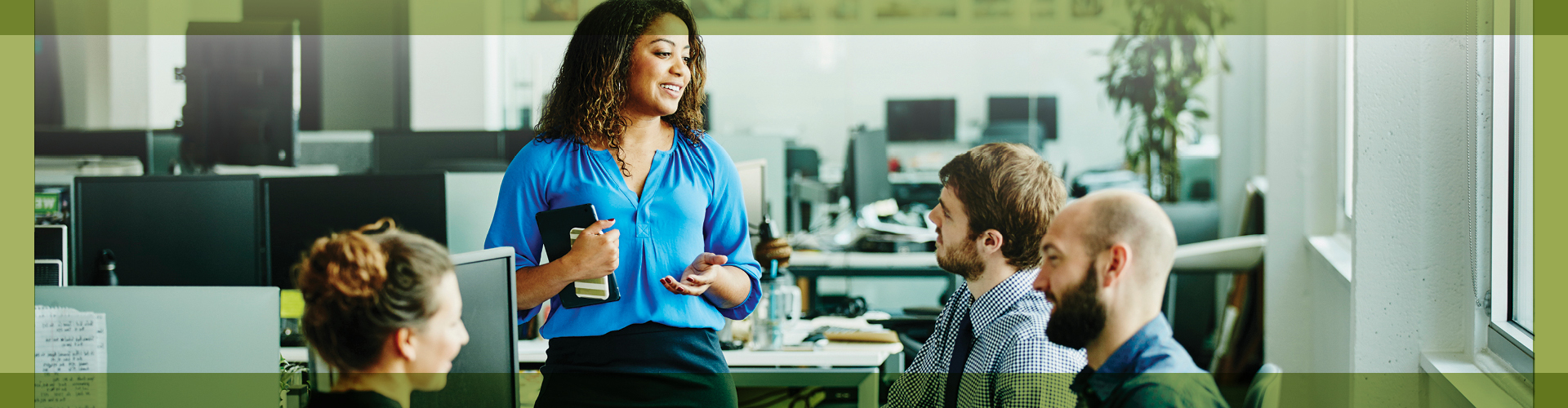 Dynamic office scene with a woman leading a discussion, confidently addressing her colleagues who are attentively listening. The environment is bustling with various workstations in the background, illustrating a vibrant and collaborative workspace.