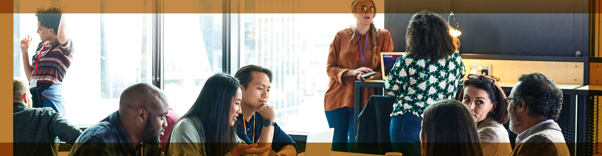 Busy workshop scene in a bright room with large windows, showing diverse participants engaged in various activities: some listen intently at a roundtable discussion, while others converse by the windows, emphasizing a dynamic learning or collaborative environment.