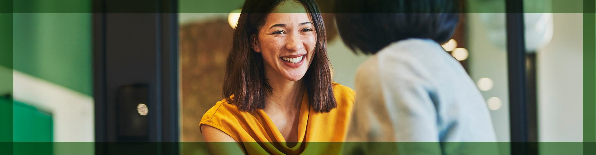 Joyful conversation between two colleagues in a bright, modern office environment, featuring a woman in a vibrant yellow top smiling warmly at her peer, emphasizing a positive and engaging workplace atmosphere.